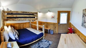 Ein Stockbett mit blauer Matratze, einem Holzkopfstück und einem kleinen Tisch mit einem Buch. Außerdem eine hölzerne Bank, ein gemustertes Teppich und eine Tür mit Fenster.