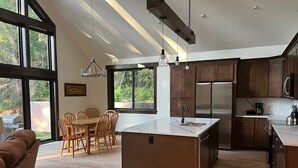 Une cuisine moderne dotée d’un îlot central, d’une table à manger en bois et de chaises, de grandes fenêtres et d’une vue sur la verdure.