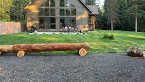 Une cabane en rondins moderne, dotée de grandes fenêtres, d’un patio avec mobilier d’extérieur et surplombant une forêt.