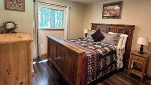 A bedroom with a wooden bed, a nightstand, a dresser, a window with curtains, and a ceiling light.
