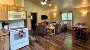 A spacious living area with a kitchen, wooden cabinets, a ceiling fan, a television, and a window with curtains.