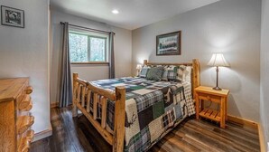 A bedroom with a wooden bed, a window with curtains, a bedside lamp, and a framed picture on the wall.