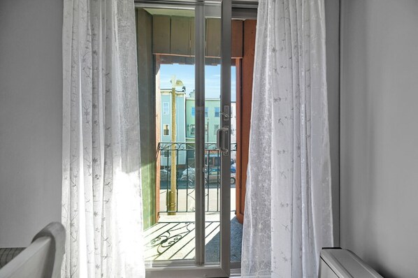 A view through a sliding glass door with white curtains, leading to a balcony with a metal railing and a small table. - 2-bedroom, 1 sofa bed next to Airport Subway, Refurbished, 1 stop to downtown (Boston)