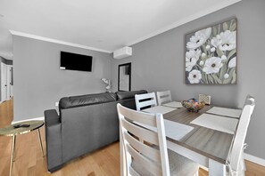 A modern living room with a grey sofa, a dining table with white chairs, and a painting of white flowers on the wall.