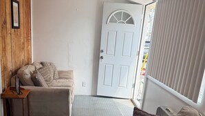 A living room with a beige sofa, a wooden sideboard, and a patterned cushion. A white door with a glass panel is visible in the background.