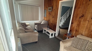 A living room with a beige sofa, patterned armchair, and a small table with a lantern. A wooden wall panel and a staircase are visible in the background.