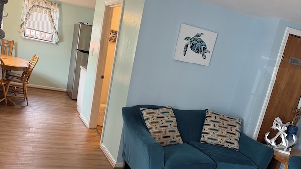 A living room with a teal sofa, wooden flooring, a dining area with a table and chairs, a kitchen with a refrigerator, and a ceiling fan.