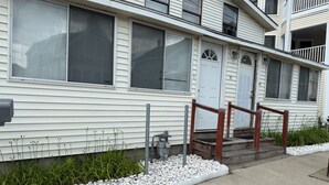 A two-story residential building with a white exterior, a front porch with a wooden railing, and a small garden area with white pebbles and green plants.