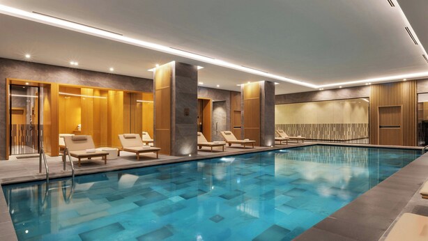 Una piscina interior moderna con agua azul cristalina, rodeada de sillones de descanso y paneles de madera.