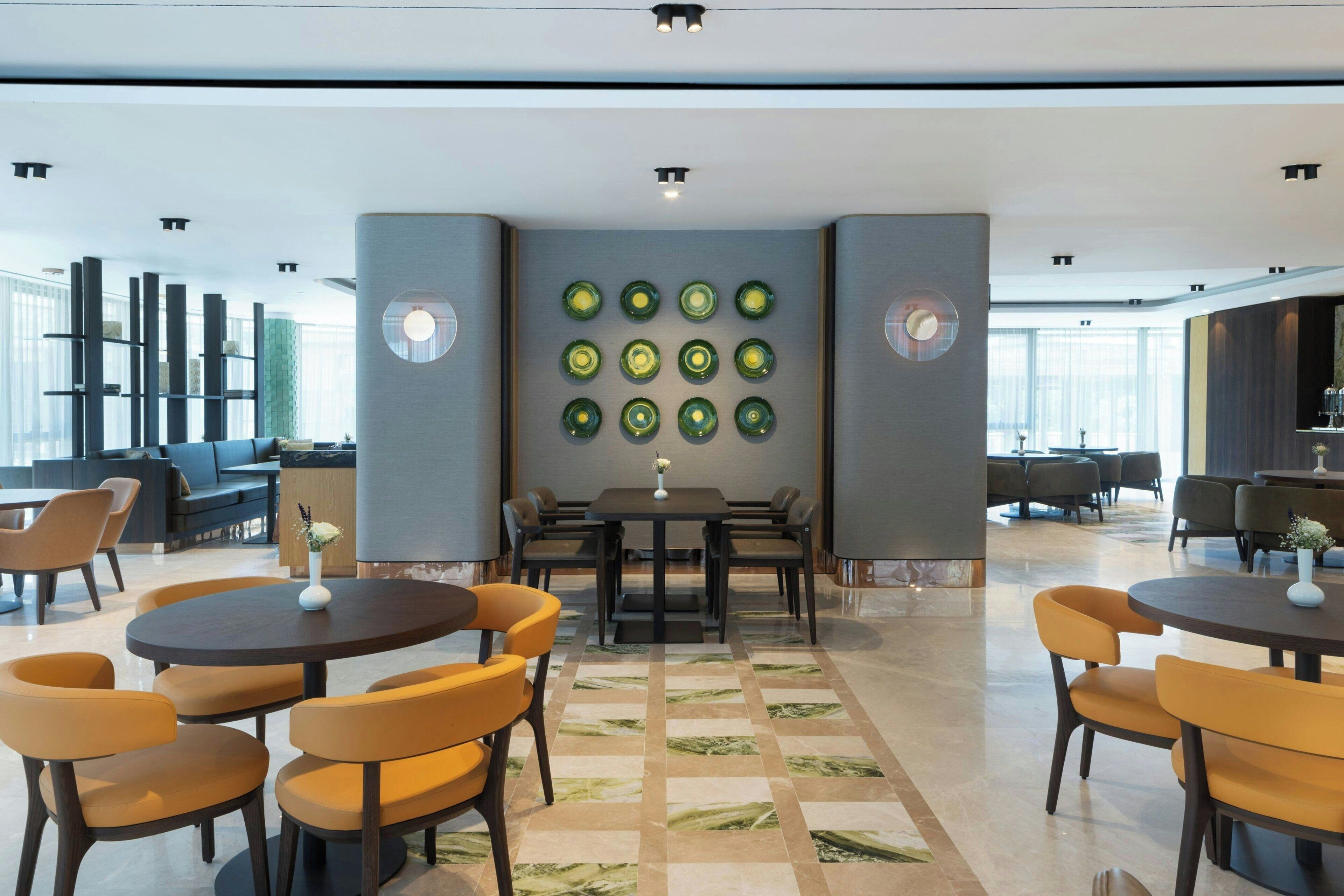 Een moderne hotel lobby met ronde tafels en stoelen, een decoratieve wand met cirkelvormige patronen, en een zithoek met banken en stoelen.