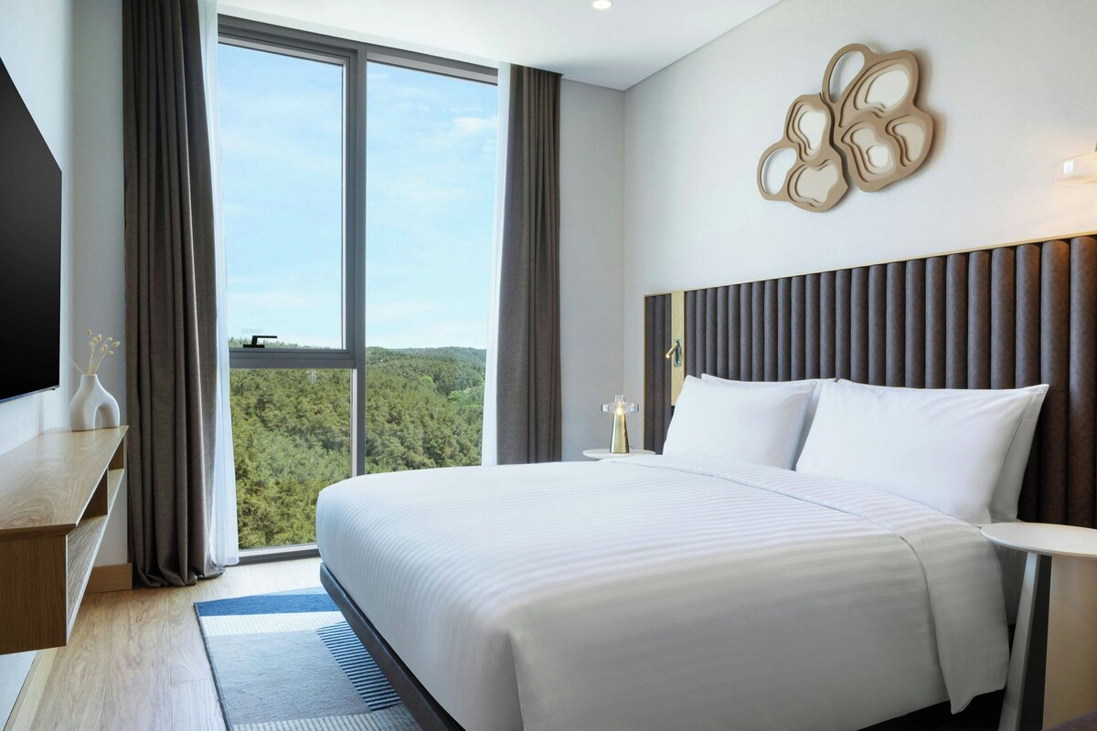 A modern hotel room with a large bed, a flat-screen TV, a desk, and a view of the forest through the window.