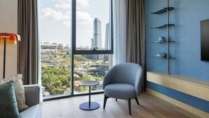 Habitación de hotel moderna con un ventanal amplio que ofrece vistas a la ciudad, un sillón gris, una pequeña mesa redonda y una unidad de estanterías empotrada.