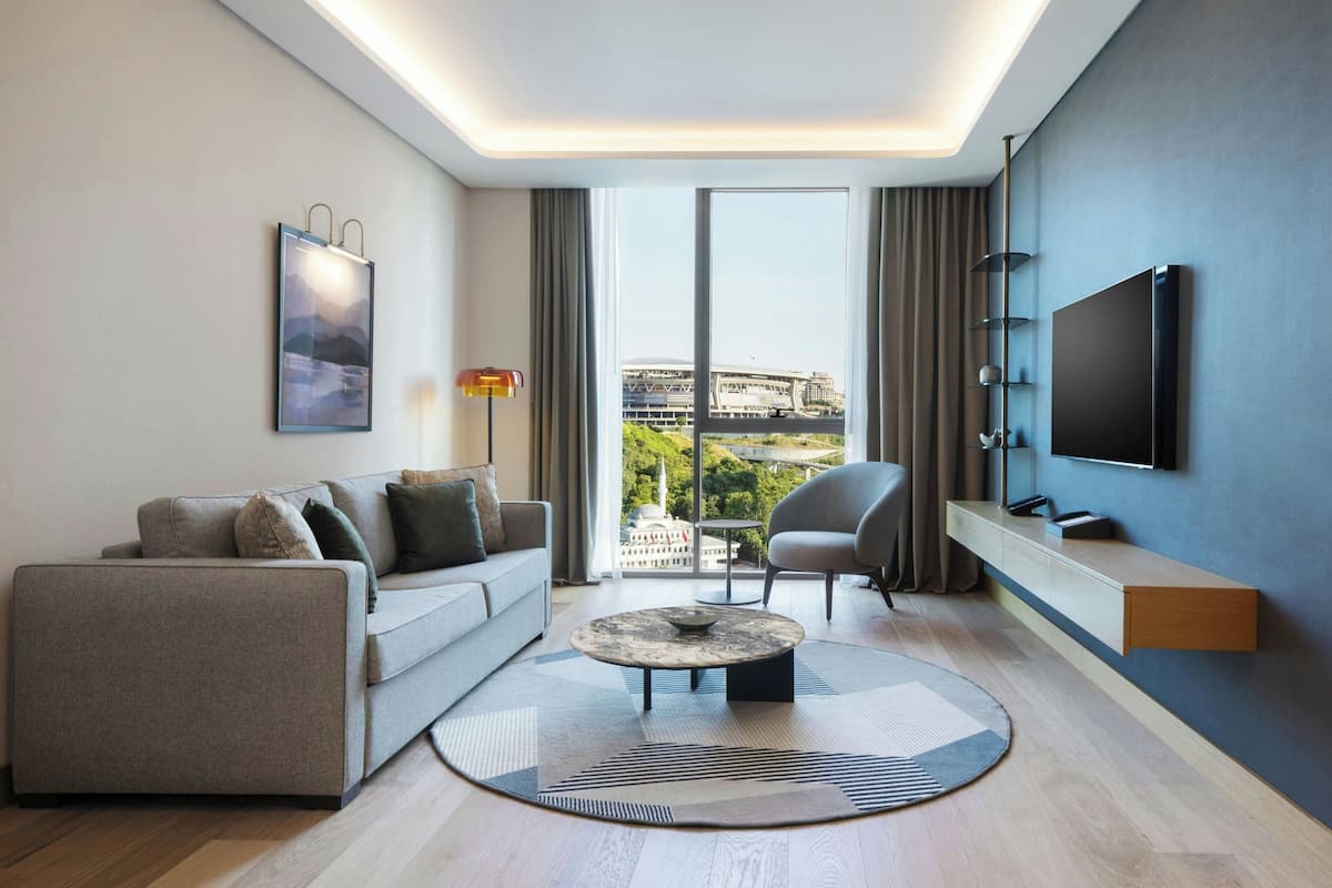 A modern living room with a sofa, armchair, and coffee table, a flat-screen TV, and a large window with a view of a building.