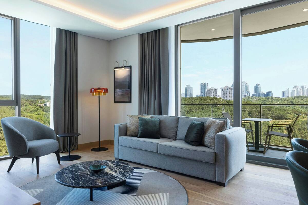 A modern living room with a grey sofa, a round coffee table, and a floor lamp. There is a large window offering a city view.