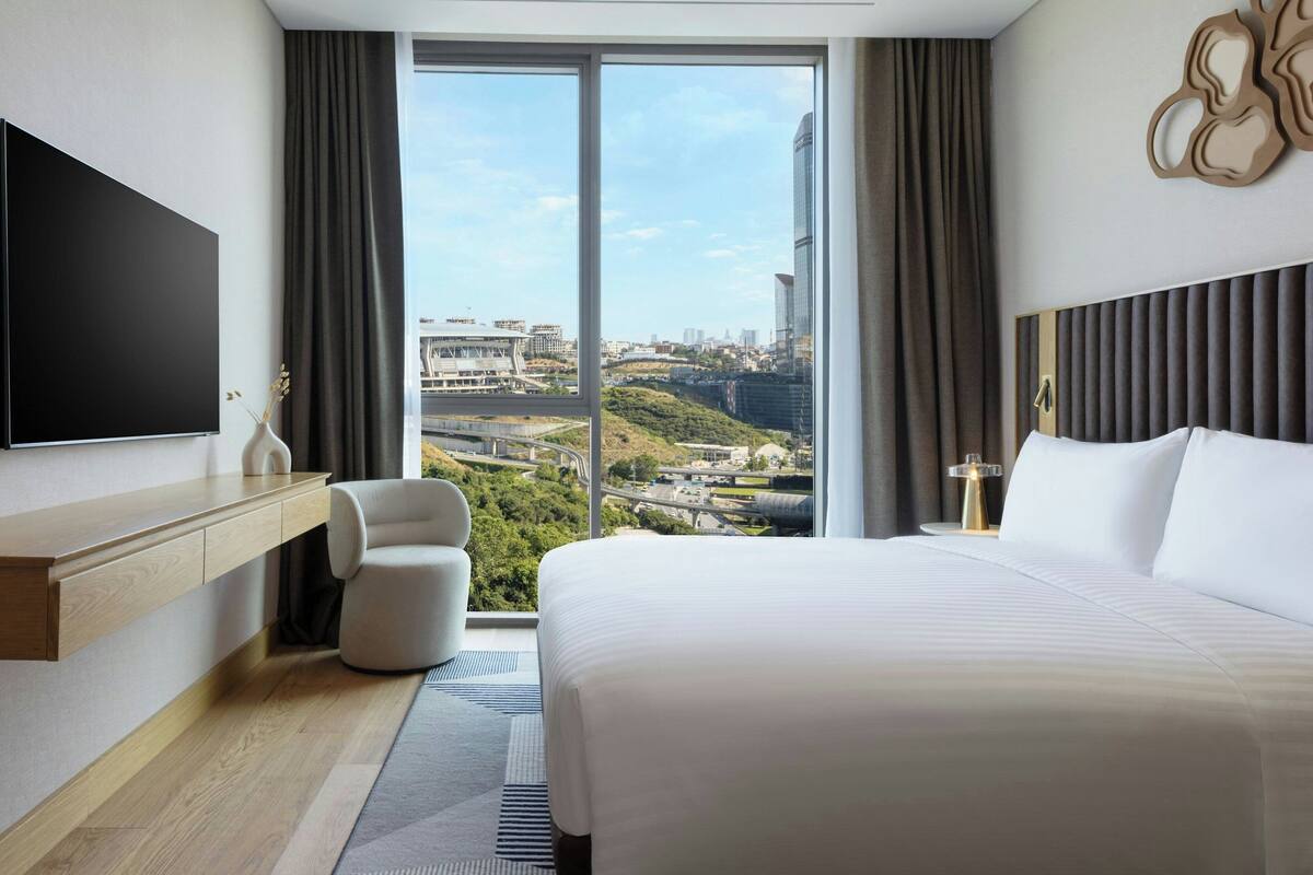 A modern hotel room with a large bed, a flat-screen TV, a seating area, and a city view.