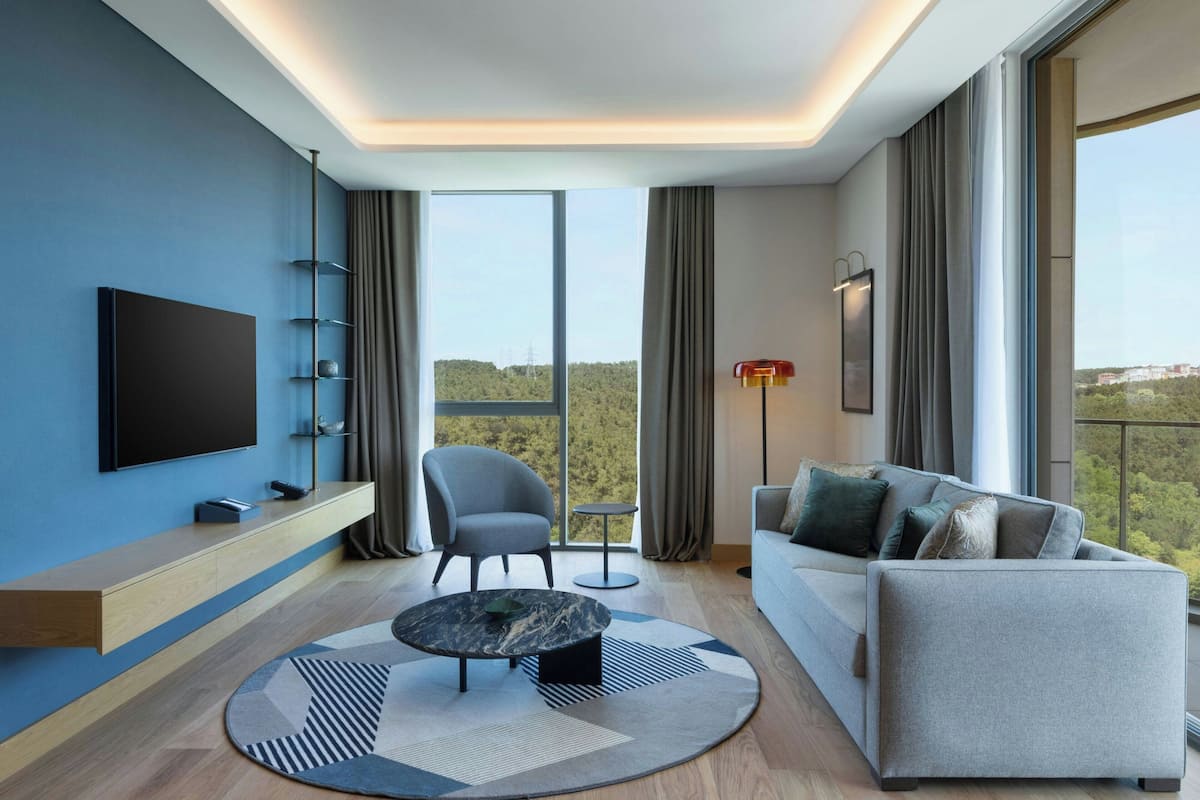 A modern living room with a flat-screen TV, a grey sofa, a round coffee table, and a large window offering a view of greenery.