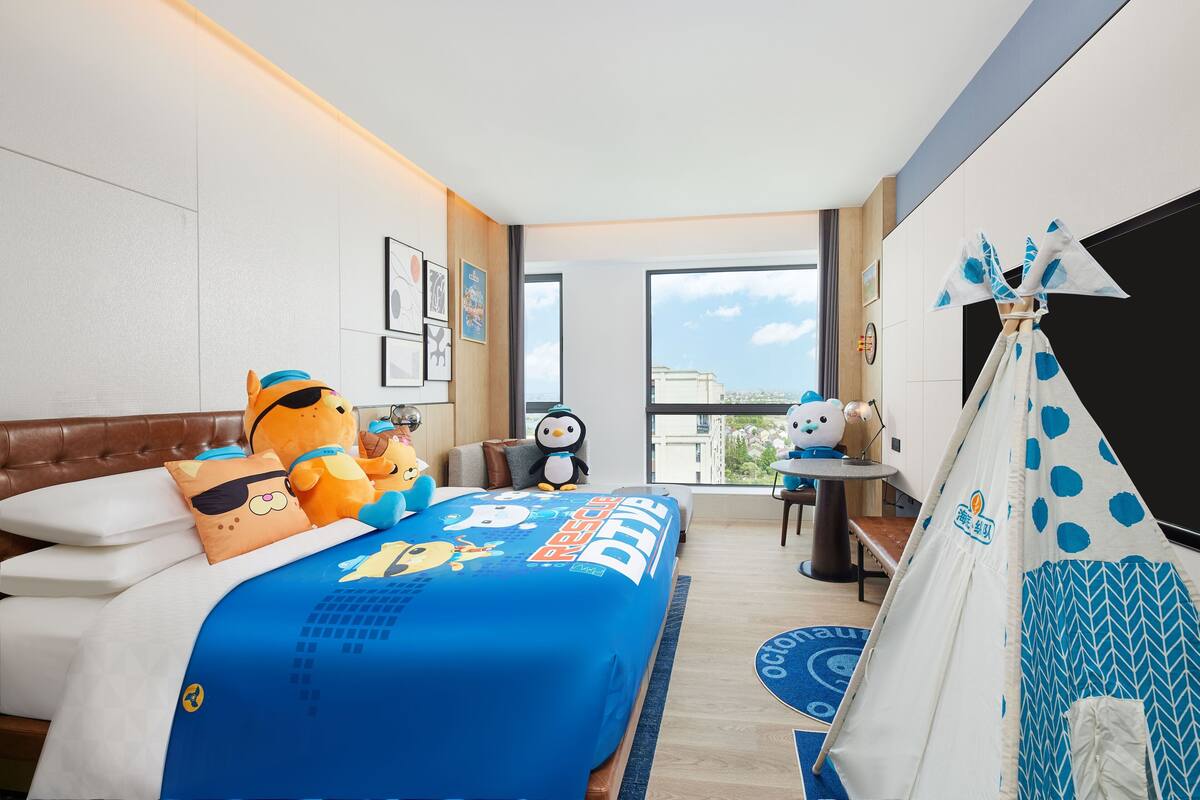 A hotel room with a bed, a tent, and a window with a city view.