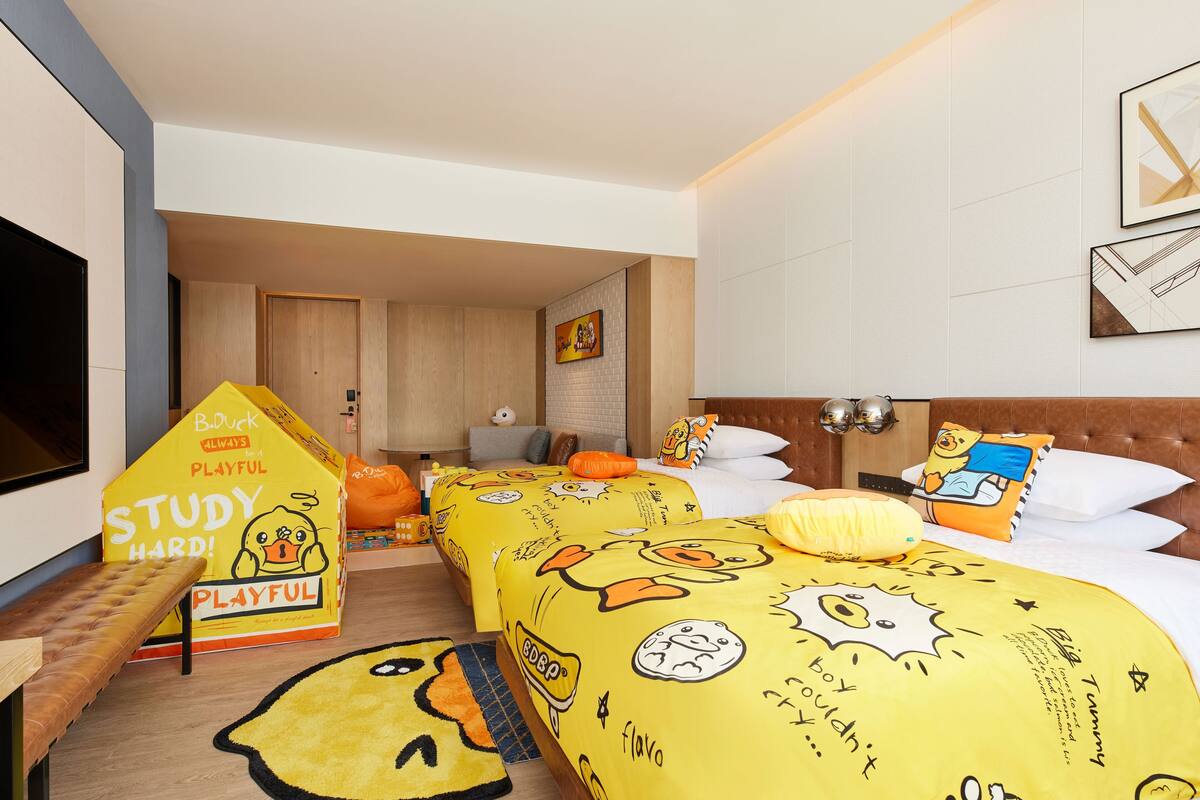 A hotel room with two beds, each with yellow bedding featuring cartoon characters. There is a study area with a desk and chair, and a large window providing natural light.