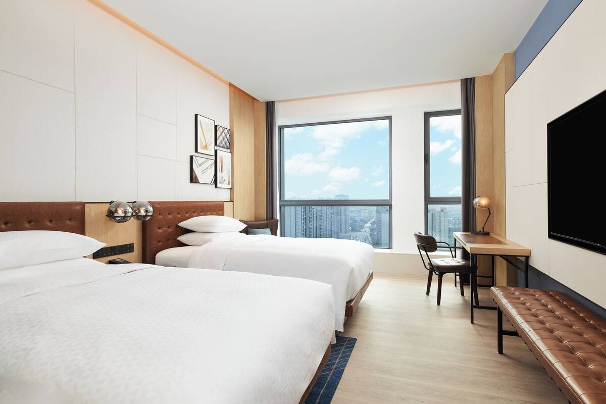 A hotel room with two beds, a desk, a chair, a bench, and a large TV.