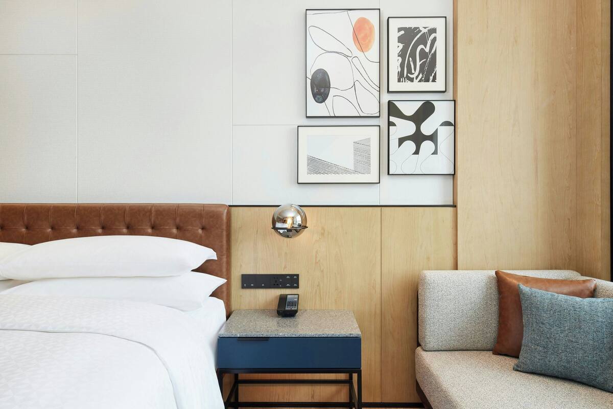 A hotel room with a bed, a small desk with a phone, and a sofa. There are framed artworks on the wall.