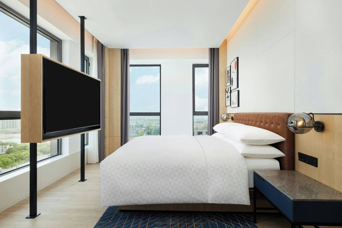 A modern hotel room with a large bed, a flat-screen TV mounted on the wall, and large windows offering a city view.