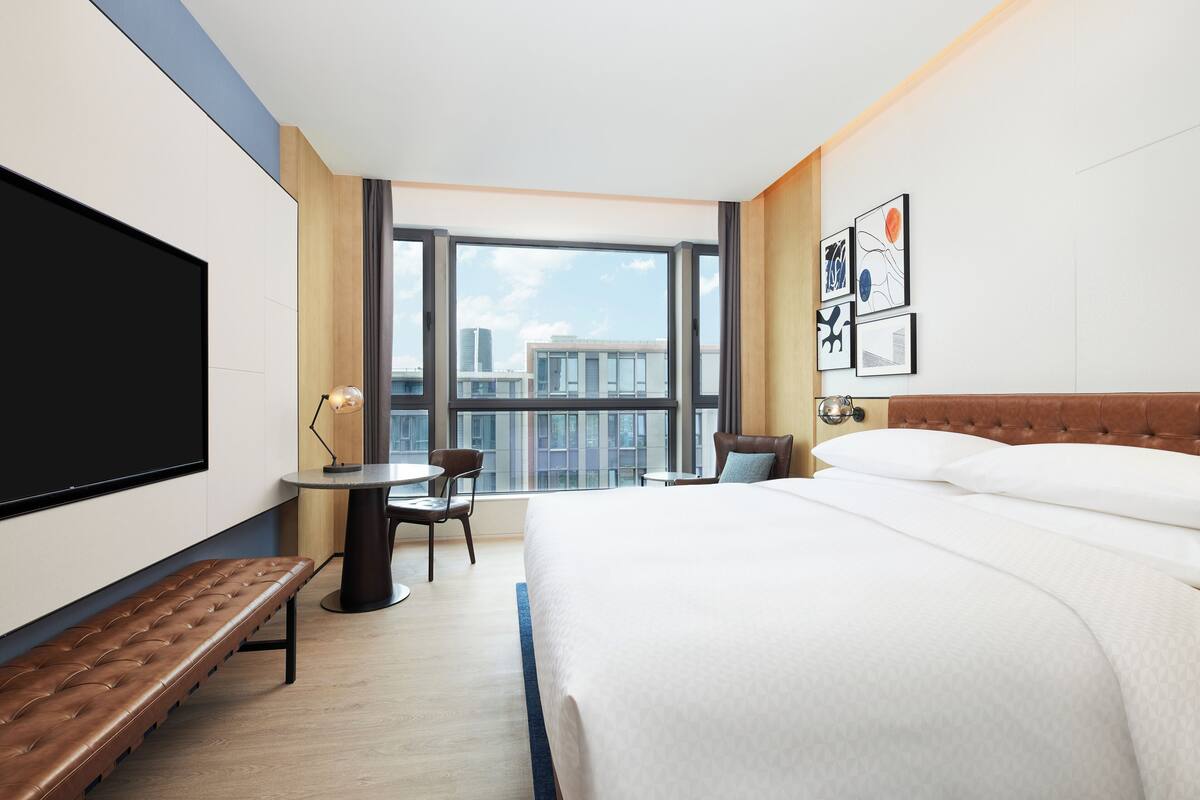 A hotel room with a large bed, a desk with a chair, a bench, a TV, and a city view through large windows.