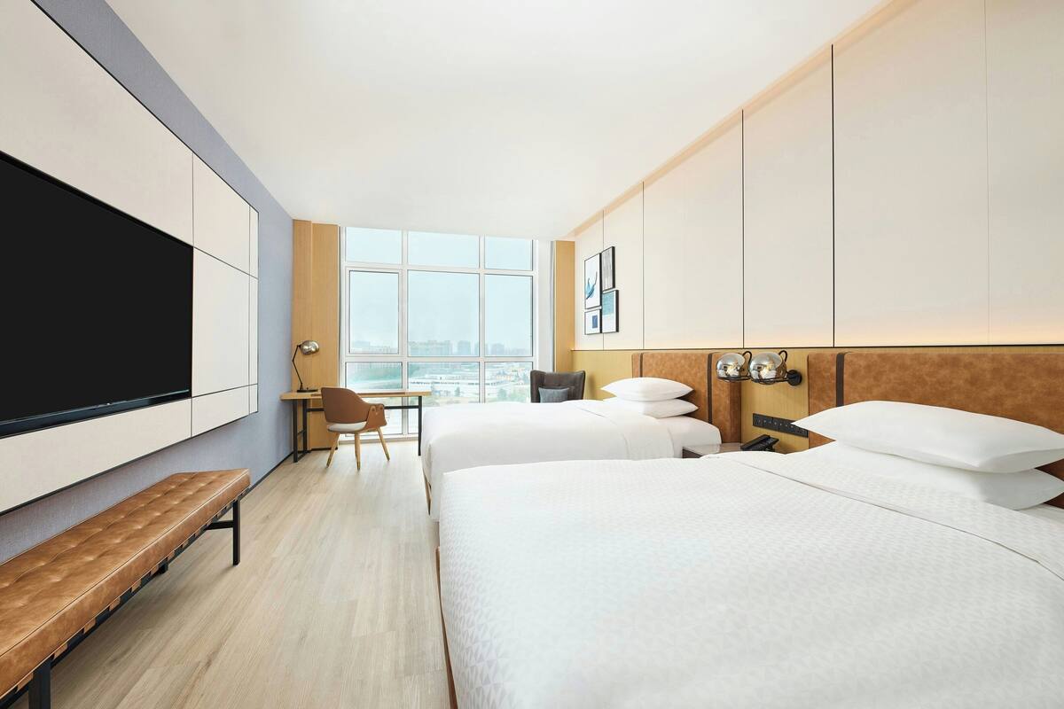 A hotel room with two beds, a desk, a chair, a bench, a TV, and a large window with a city view.