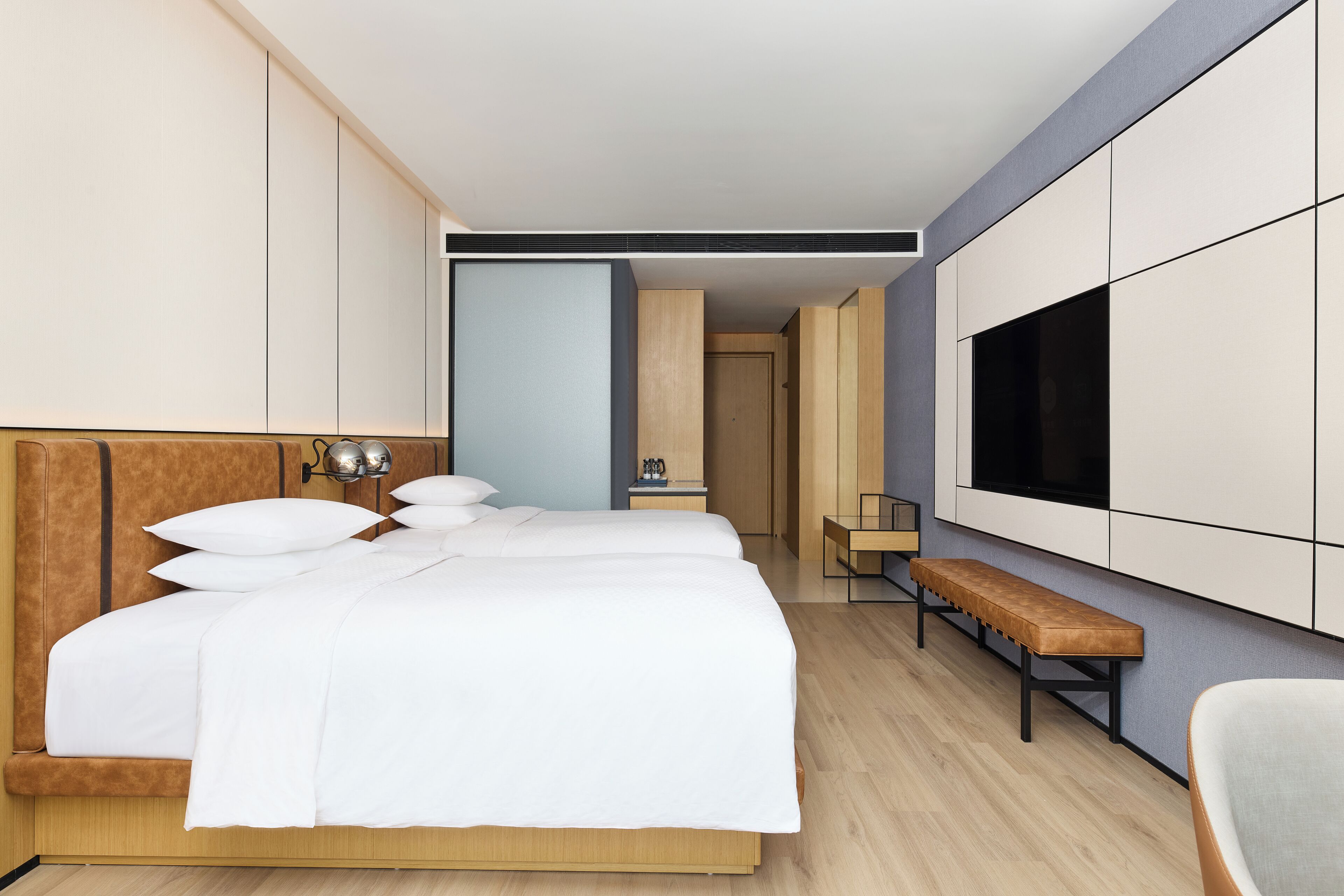 A modern hotel room with a large bed, a desk, a bench, and a TV mounted on the wall.