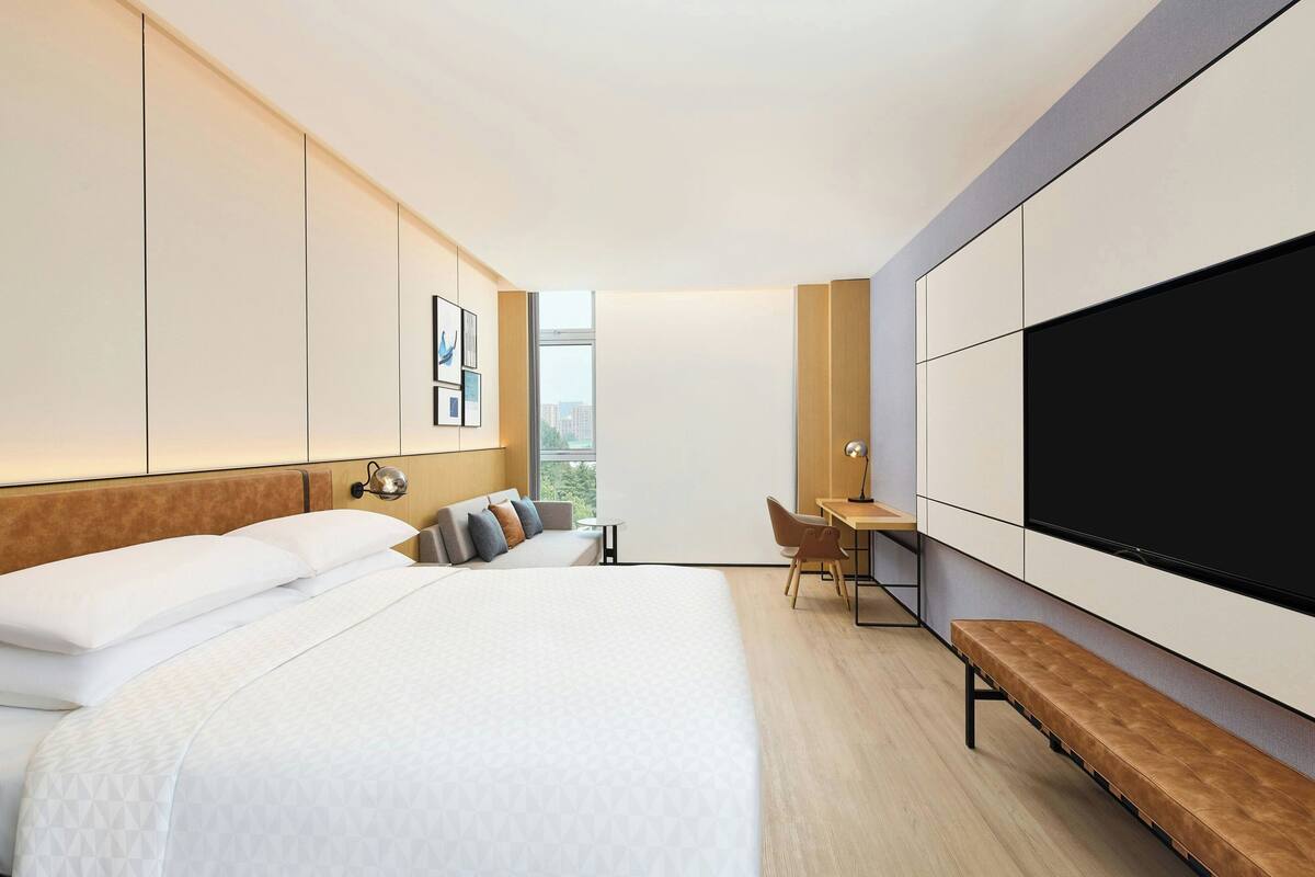 A modern hotel room with a large bed, a desk, a bench, and a TV.
