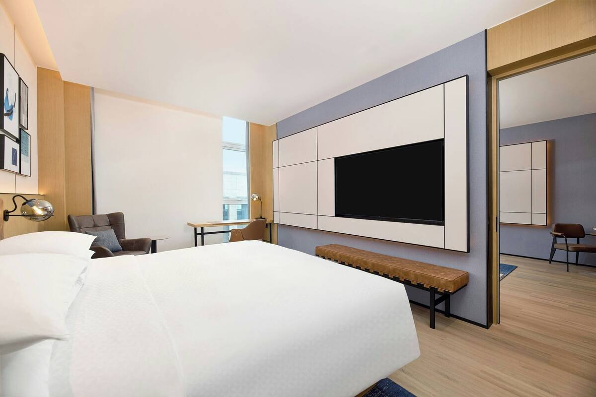 A modern hotel room with a large bed, a flat-screen TV mounted on the wall, a desk with a chair, and a bench.