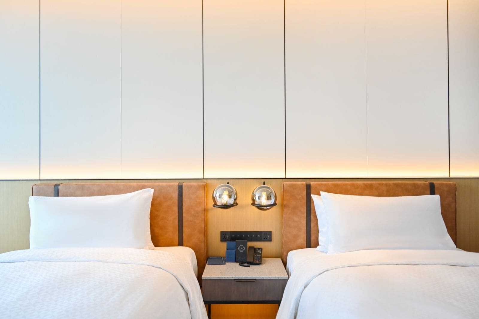 Habitación de hotel con dos camas, una mesita de noche con teléfono y lámpara, y una lámpara de pared.