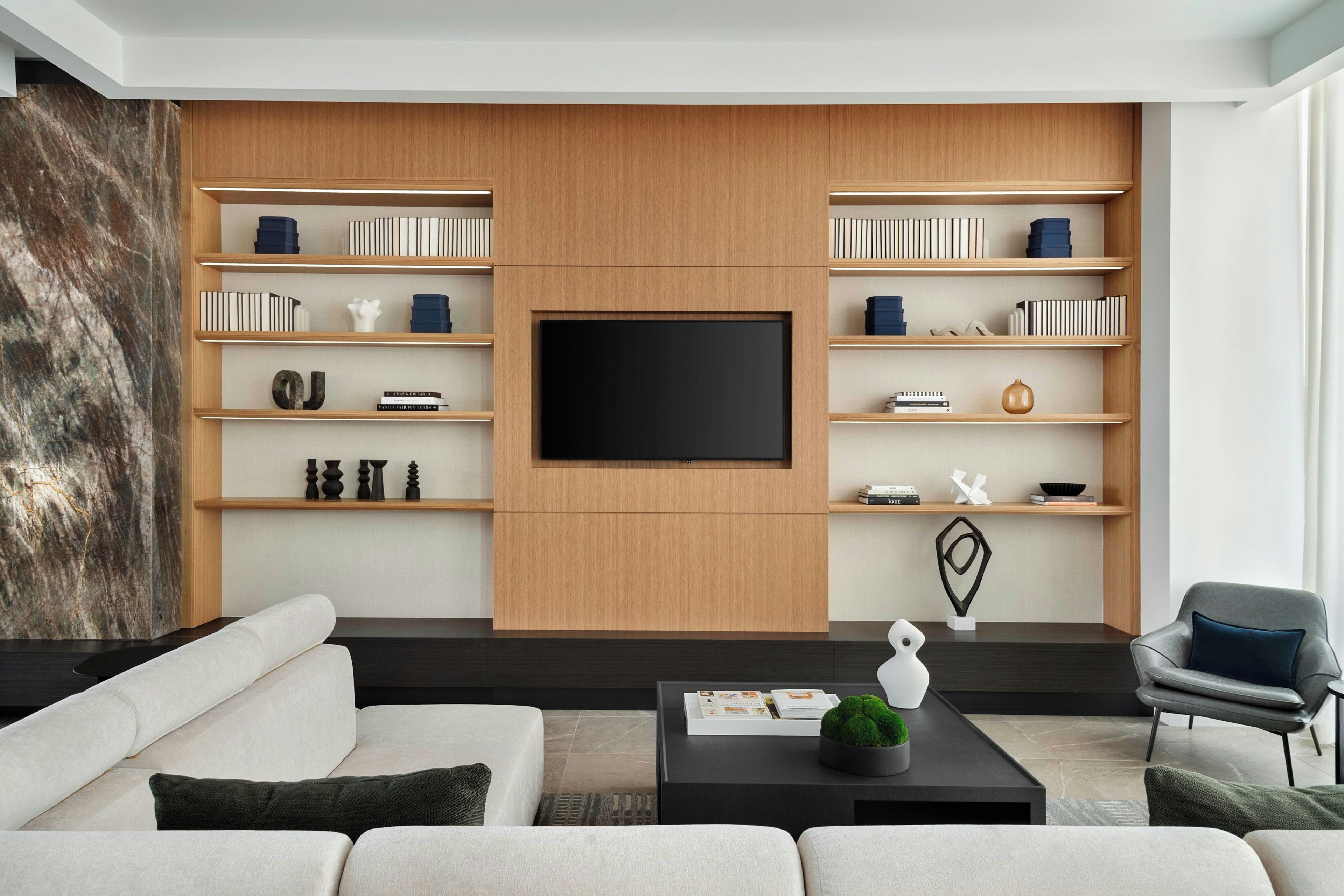 A modern living room with a large flat-screen TV, built-in wooden shelving with books and decorative items, a black coffee table with a plant, and a grey armchair.