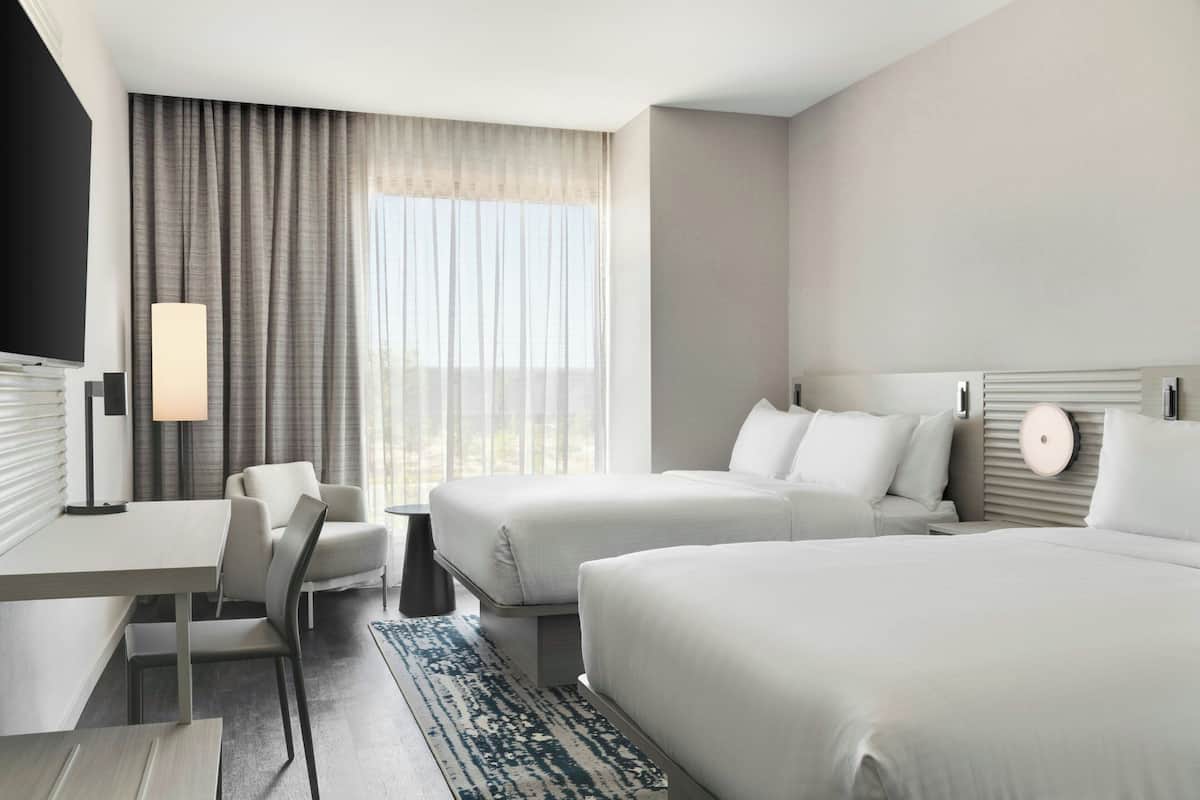 Quarto de hotel com duas camas, uma escrivaninha, uma cadeira, um abajur e vista para o exterior.