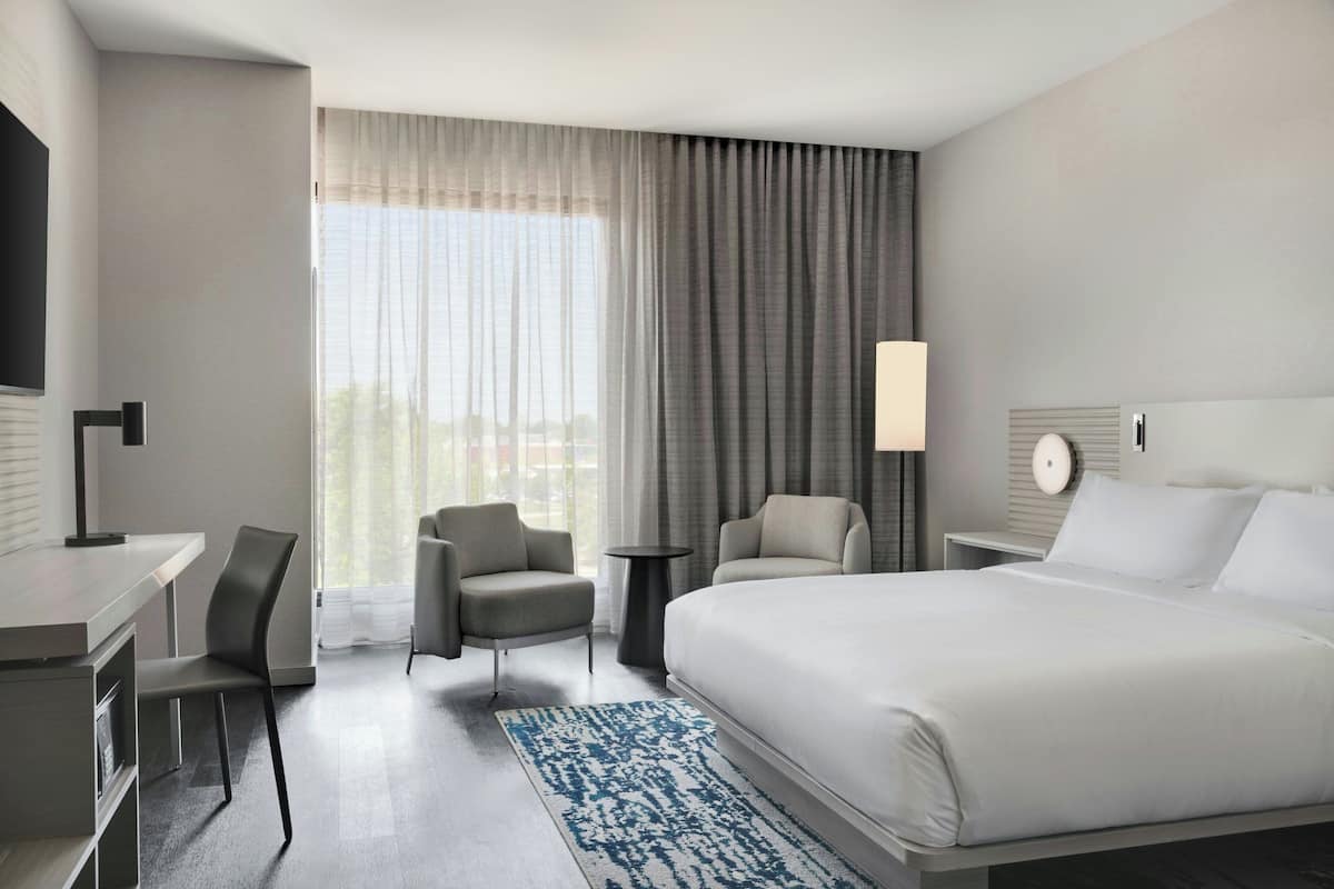 Quarto de hotel moderno com uma cama grande, uma escrivaninha, duas cadeiras, uma mesa pequena e uma TV.