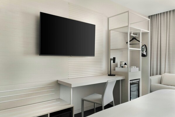 A modern hotel room with a flat-screen TV mounted on the wall, a desk with a chair, a sofa, and a wardrobe with a mirror.