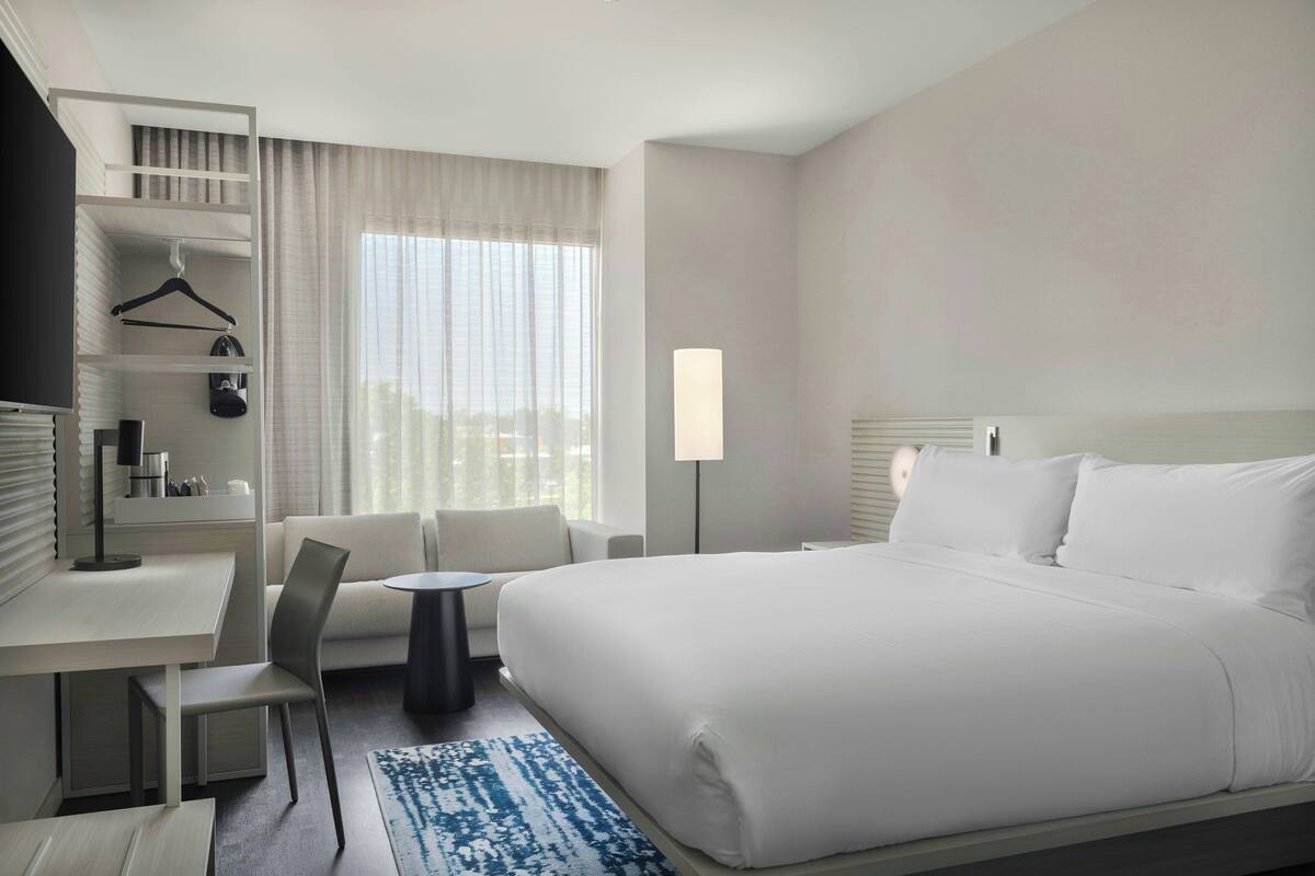 Quarto de hotel com uma cama grande, uma escrivaninha com cadeira, uma mesinha pequena, um cabideiro e uma janela ampla com cortinas.