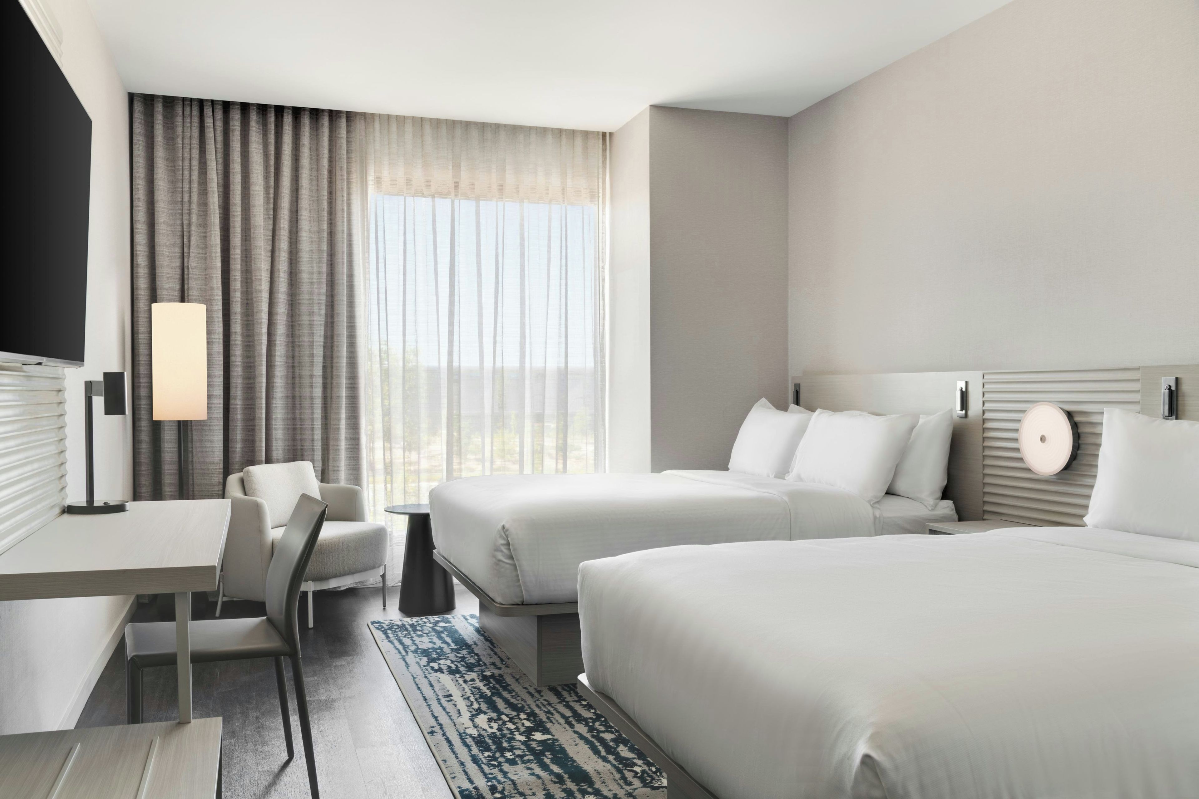 A hotel room with two beds, a desk, a chair, a lamp, and a view of the outdoors.