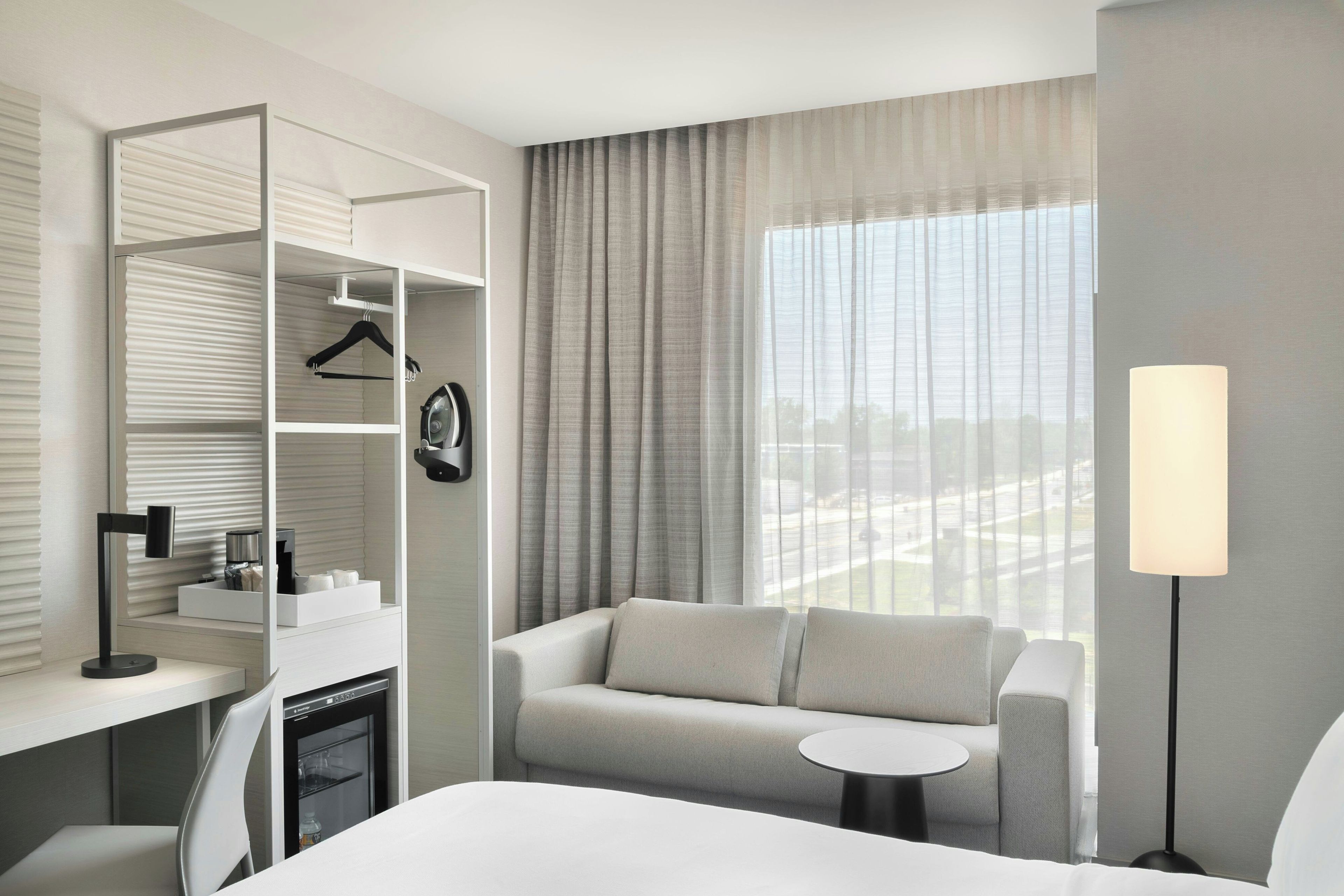 Habitación de hotel con cama, sofá, una mesita, escritorio con lámpara, minibar y armario con perchero.
