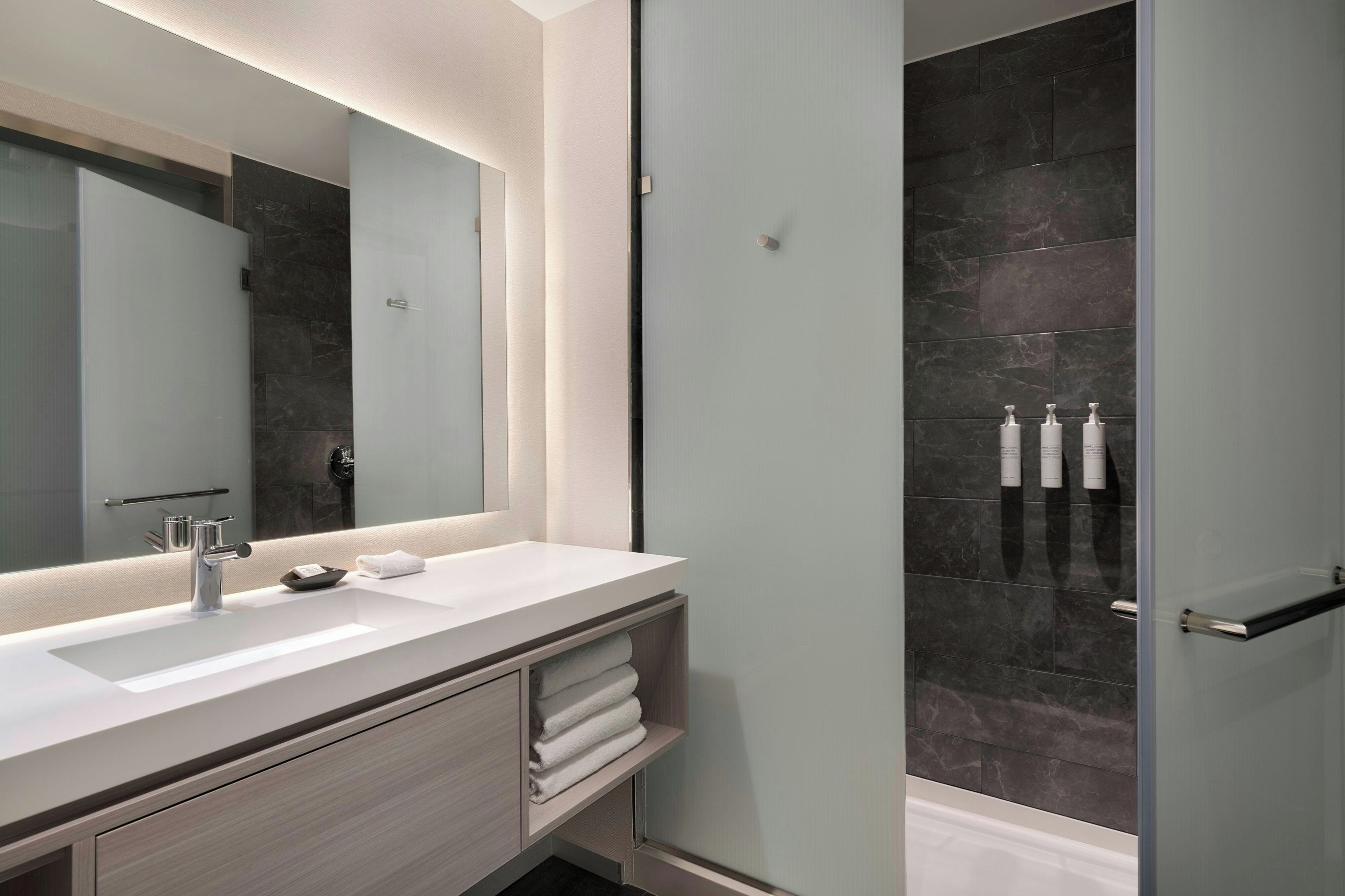 A modern bathroom with a large mirror, white sink, and dark tiled shower area.