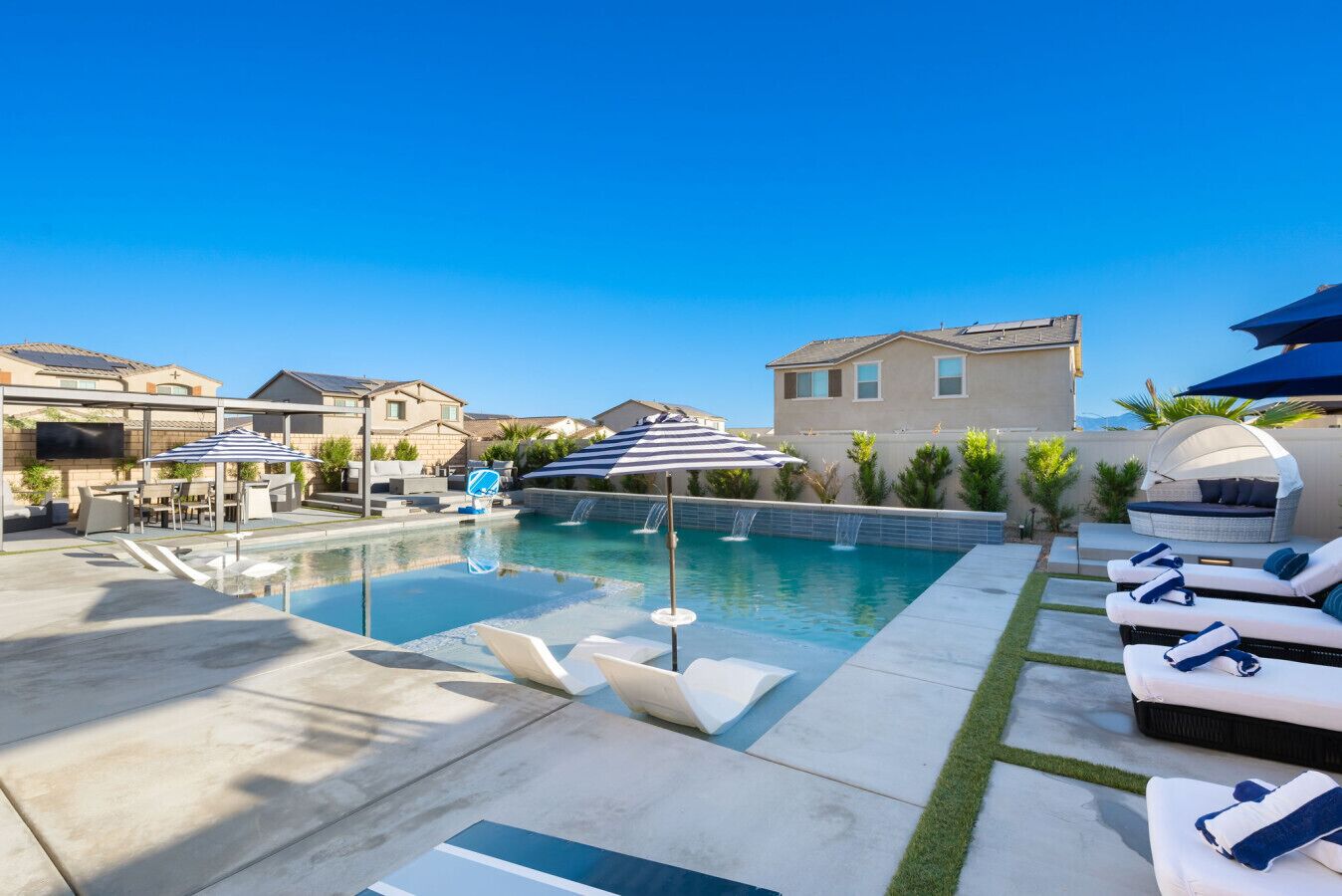 Ein Swimmingpool mit Liegestühlen und Sonnenschirmen, umgeben von Häusern und einem klaren Himmel.