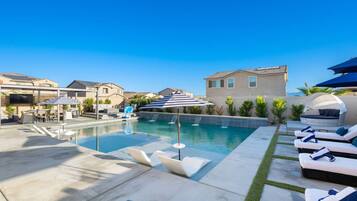 Ein Swimmingpool mit Liegestühlen und Sonnenschirmen, umgeben von Häusern und einem klaren Himmel.