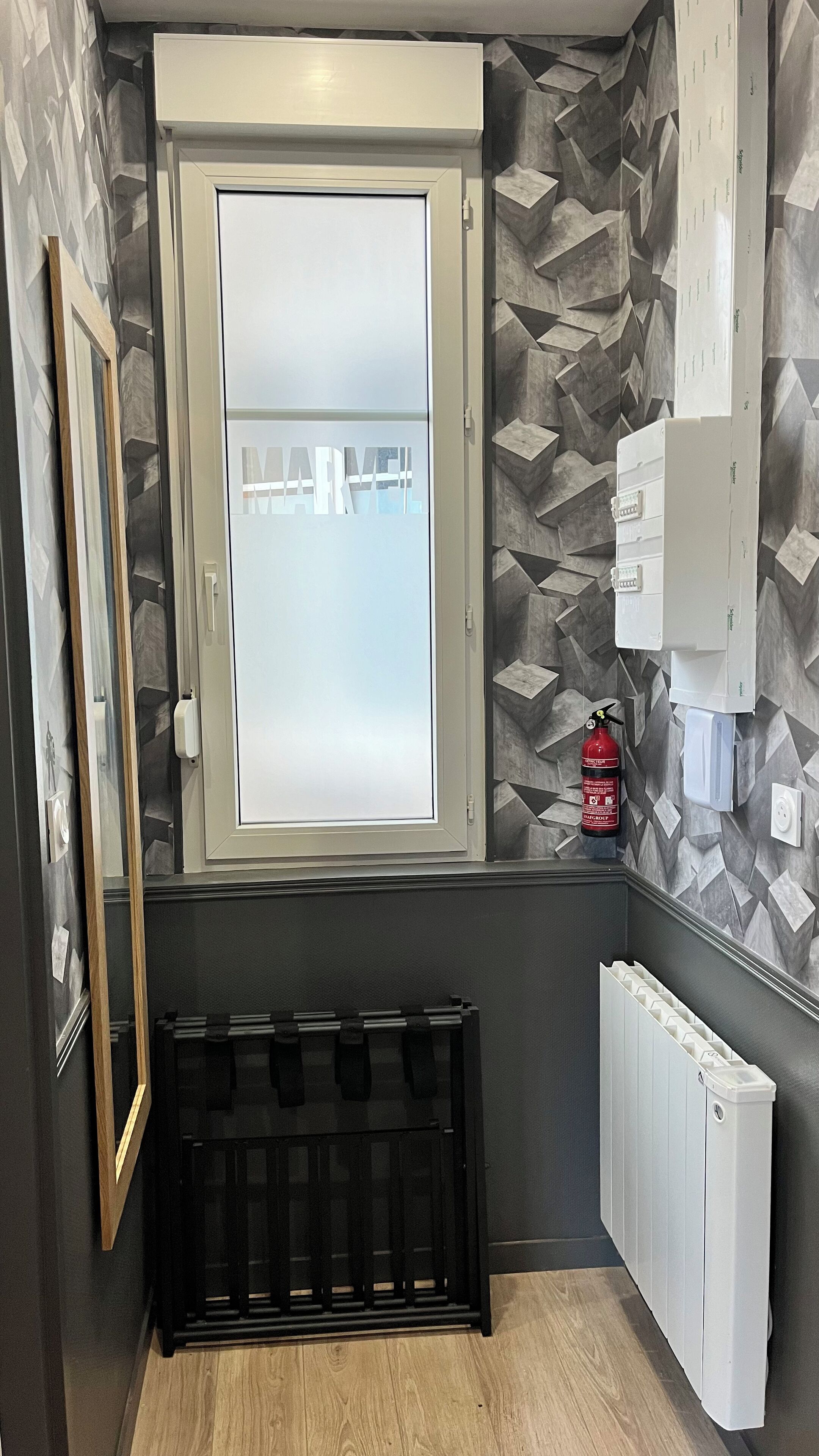 A small room with a frosted window, a radiator, a fire extinguisher, and a wall-mounted electrical box.