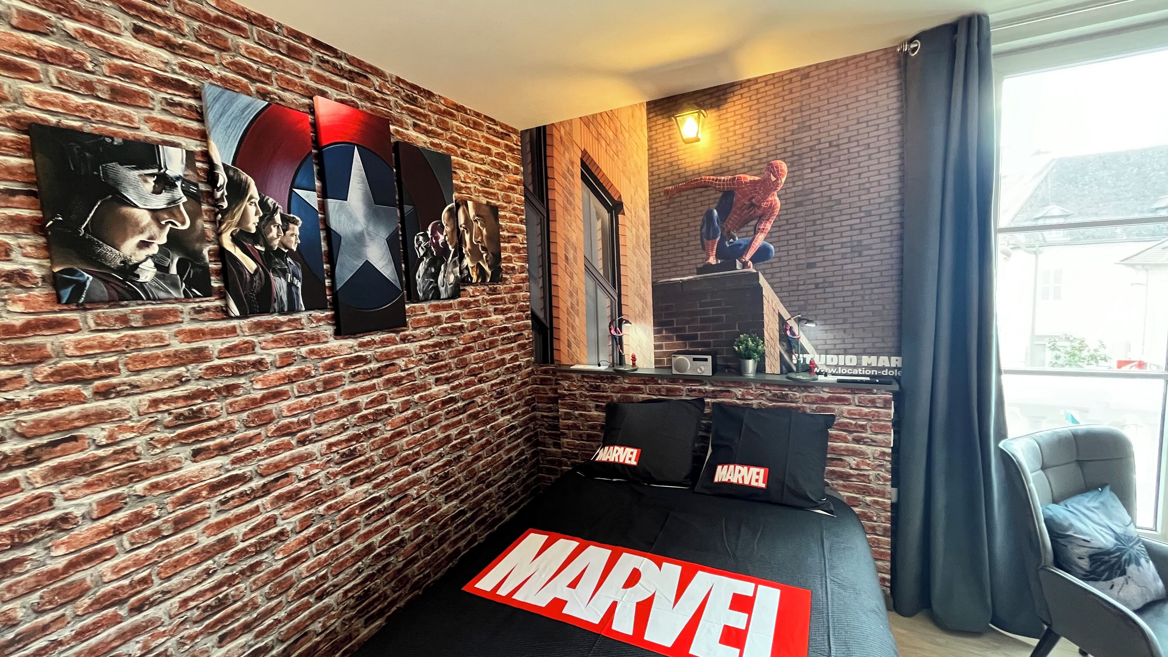 Huone, jossa on tiiliseinä, sänky, jossa on «MARVEL»-kuvioisia tyynyjä, ja Hämähäkkimies-juliste.
