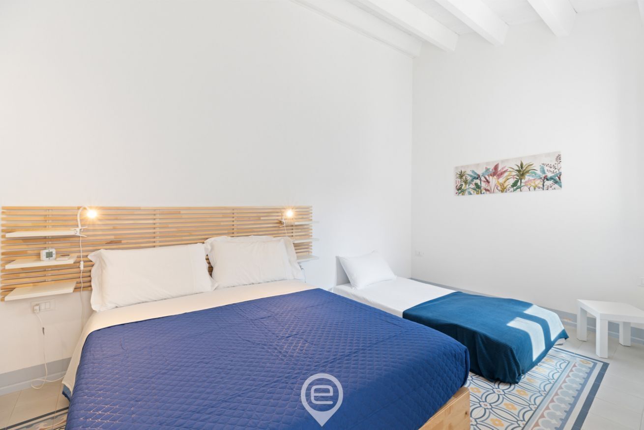 A modern hotel room with a large bed, a smaller bed, a bedside table, and a wall-mounted shelf.