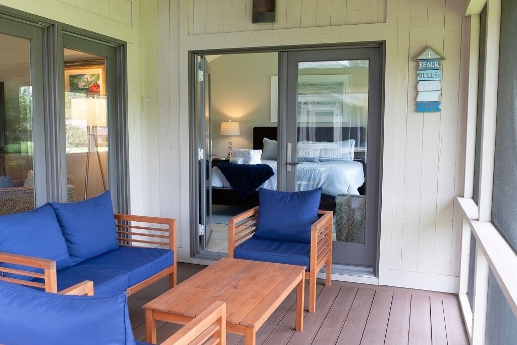 A room with a wooden floor, a sofa, a chair, and a table. There is a sliding glass door leading to a bedroom with a bed and a nightstand.