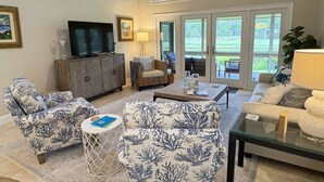 Un salon spacieux doté d’un grand téléviseur, de sièges confortables et offrant une vue sur un jardin grâce à de grandes portes vitrées.