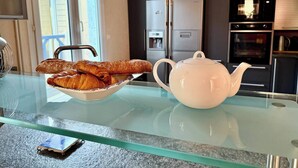 Une cuisine avec une table en verre, un bol de croissants, une théière et un réfrigérateur.