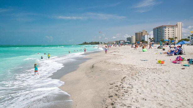 A beach with clear turquoise water, people swimming and walking along the shore, and buildings in the background.