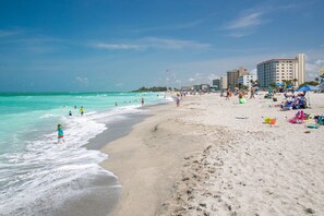 A beach with clear turquoise water, people swimming and walking along the shore, and buildings in the background.