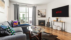 A modern living room with a grey sofa, a glass coffee table, a fireplace, a guitar, and a TV displaying the Netflix logo.